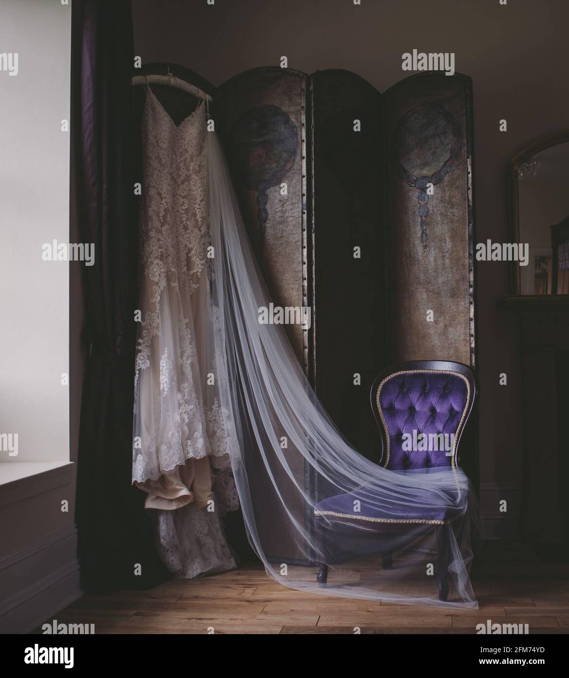 Ein Hochzeitskleid mit Spitze und ein Schleier hängen wunderbar in der Fensterbeleuchtung Stockfoto