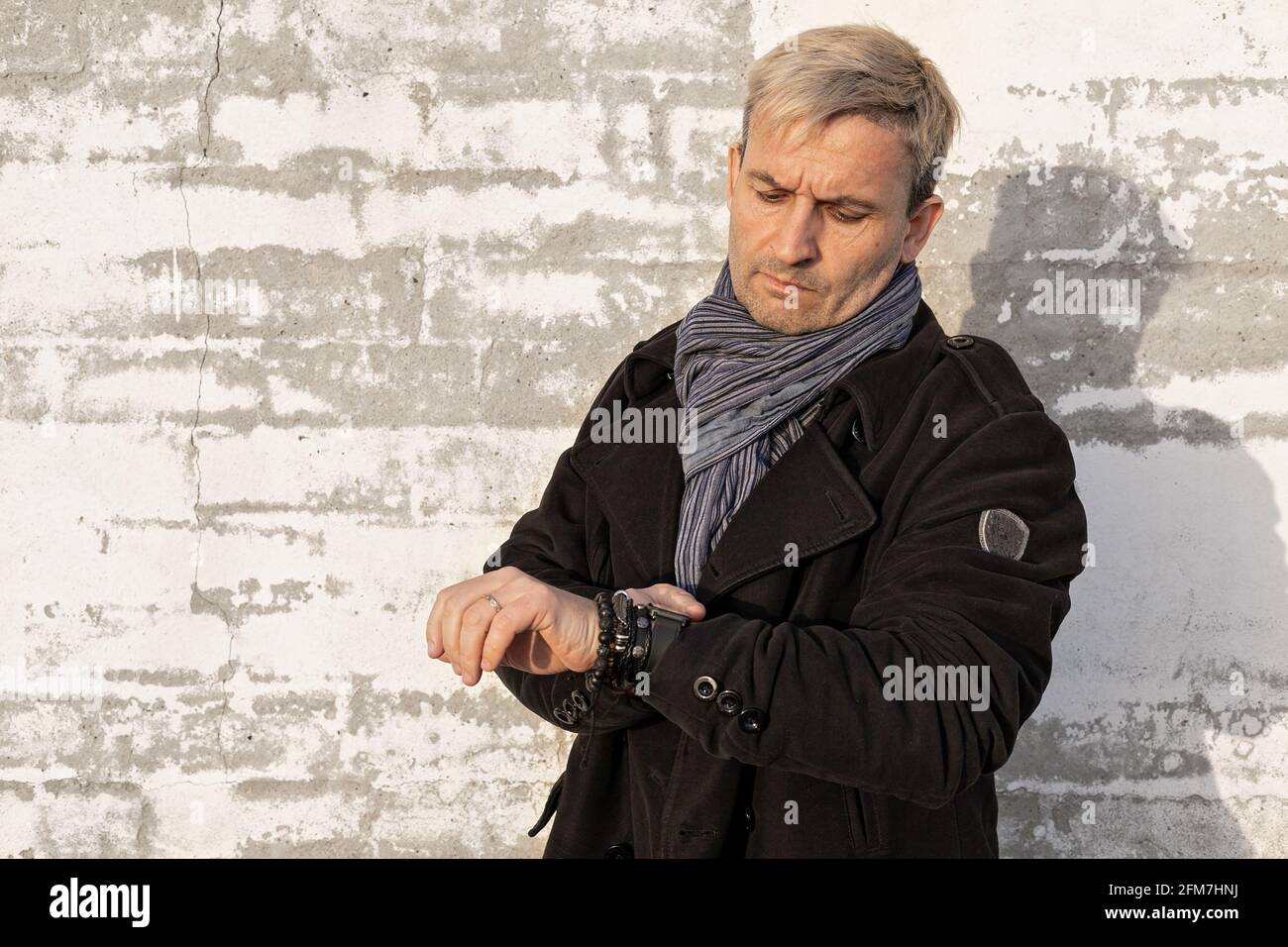 Charmanter blonder Mann mittleren Alters mit Model-Auftritt vor einer weißen Backsteinmauer. Der Mann sieht die Zeit auf seiner Armbanduhr an. Männliches Modell Stockfoto