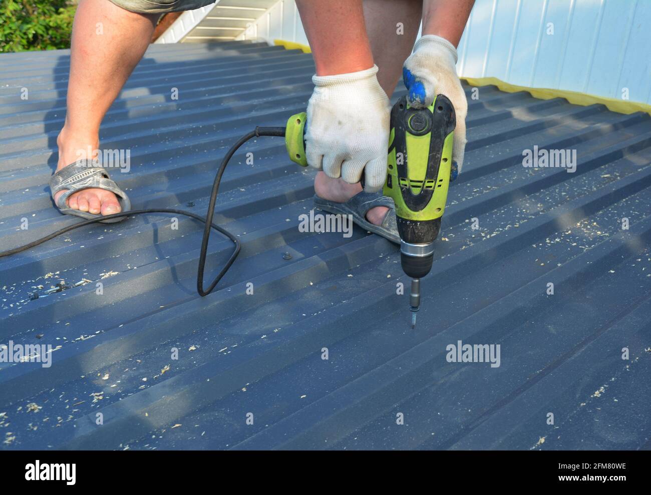 Dachkonstruktion: Ein Dachdecker installiert Dachbleche aus Metall, Dachziegel aus Metall, die Schrauben mit einem elektrischen Schraubendreher anziehen. Stockfoto