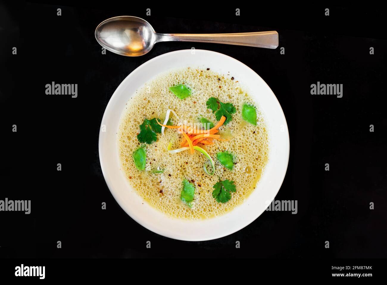 Aufgeschlagene, kräftige Spargelpüree-Suppe mit Kräuter- und Gemüsejulienne auf weißem Teller auf schwarzem Hintergrund. Stockfoto