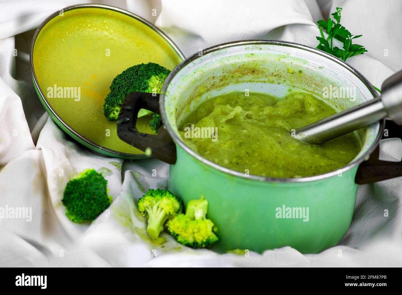 Stabmixer in grünem Topf mit Brokkoli-Suppe auf weißem Hintergrund. Zubereitung von vegetarischer Brokkoli-Suppe. Stockfoto
