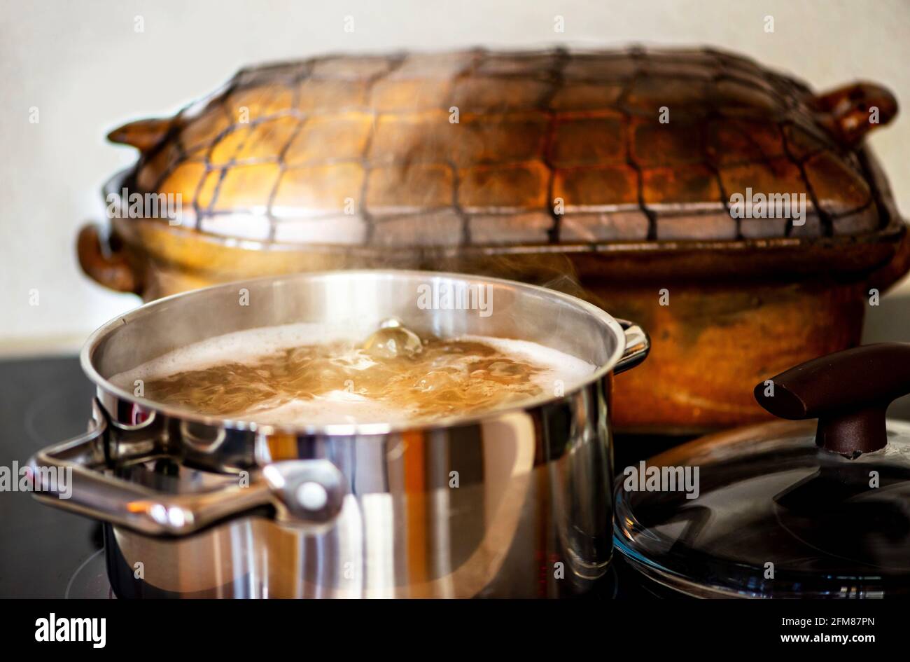 Stahltopf mit kochender sprudelnder Flüssigkeit (Brühe) und aufsteigendem Dampf auf heissem Keramikherd, dahinter alte rustikale Steingutpfanne, Deckel rechts. Stockfoto