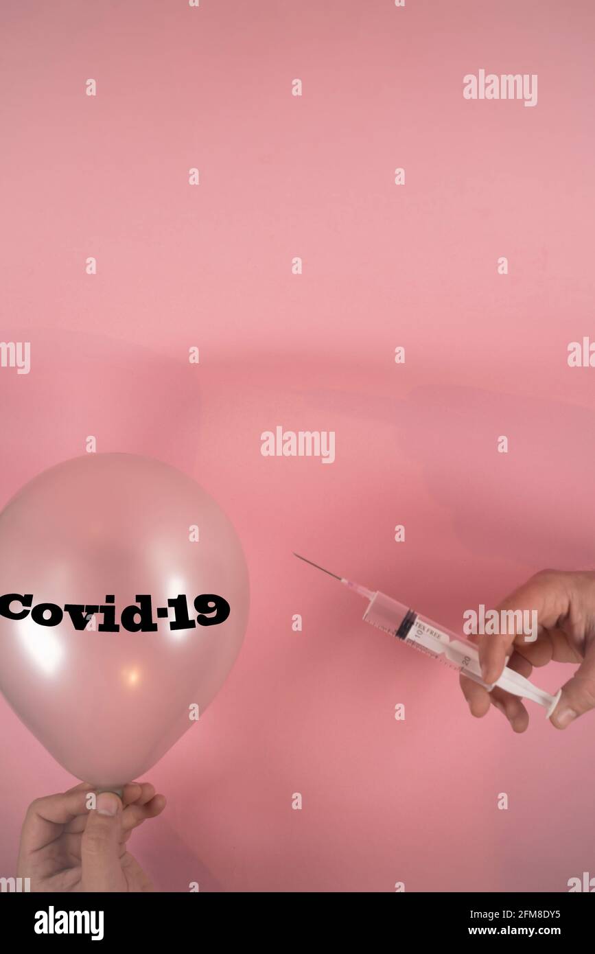 Hände mit Impfstoffspritze mit Nadel und Ballon mit Covid-19-Schrift. Auf einem weichen rosa Hintergrund. Minimales abstraktes Konzept der Coronavirus-Pandemie. Stockfoto