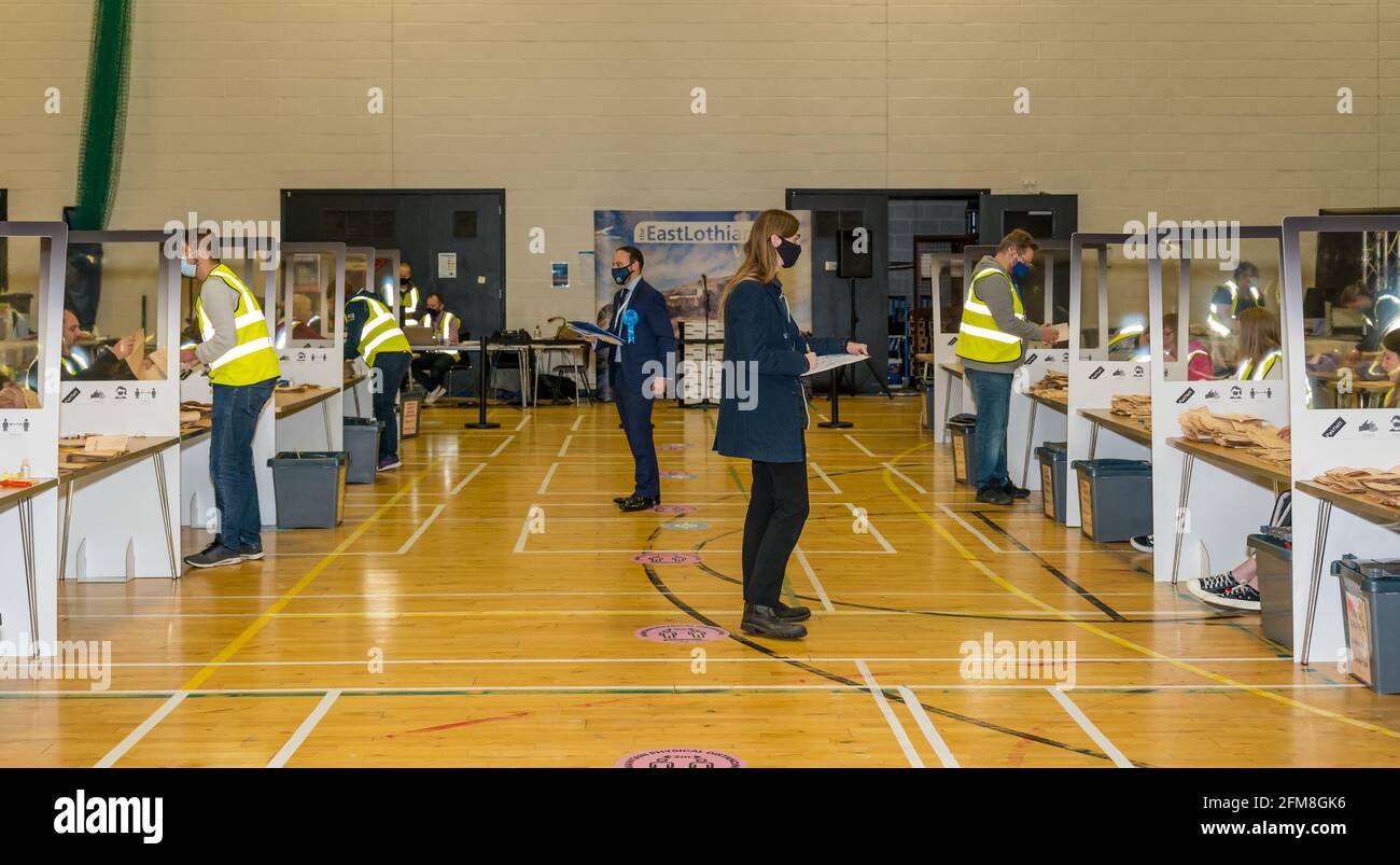 Meadowmill Sports Center, East Lothian, Schottland, Großbritannien. Mai 2021. Die Wahl zum schottischen parlament zählt für den Wahlkreis East Lothian: Mitarbeiter der Wahlhelfer sortieren die Stimmzettel und zählen die Stimmen, während die Kandidaten mit Gesichtsmasken und sozial distanziert zusehen Stockfoto