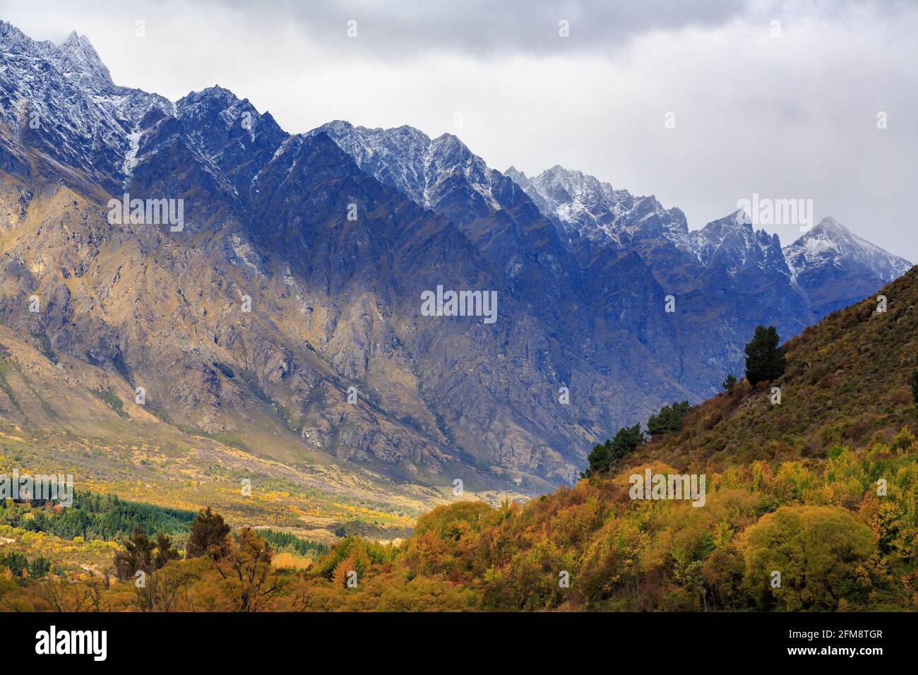 The Remarkables Mountain Range in der Nähe von Queenstown, Neuseeland, mit Schnee bestäubt. Im Vordergrund ist ein Wald mit farbenfrohem Herbstlaub Stockfoto