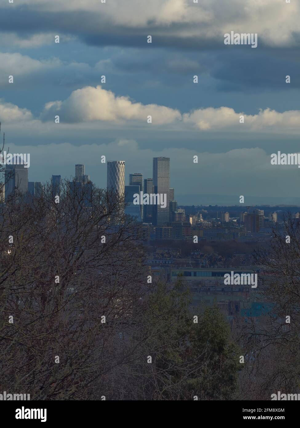 Eine Skyline von London aus dem Archway, mit Bäumen im Vordergrund und sonnenbeschienenen Wolkenkratzern am Horizont, unter einem bergigem, wolkenbedecktem Himmel. Stockfoto