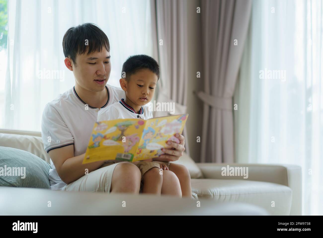 Der asiatische Vater hält ein Buch, um seinen Kindern das Lesen auf dem Sofa im Wohnzimmer beizubringen, Väter interagieren den ganzen Tag mit ihren Kindern. Stockfoto