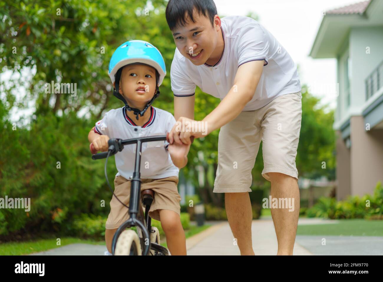 Der asiatische Vater lehrt seinem Kind, wie man in einem Nachbarschaftsgarten Fahrrad fährt, Väter interagieren mit ihren Kindern den ganzen Tag über. Stockfoto