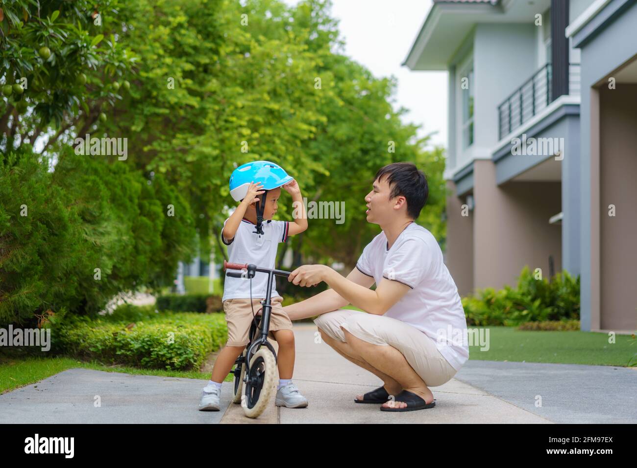Der asiatische Vater trägt einen Helm für seinen Sohn während er seinem Kind beibringt, wie man in einem Nachbarschaftsgarten Fahrrad fährt, interagieren Väter mit ihren Kindern Stockfoto