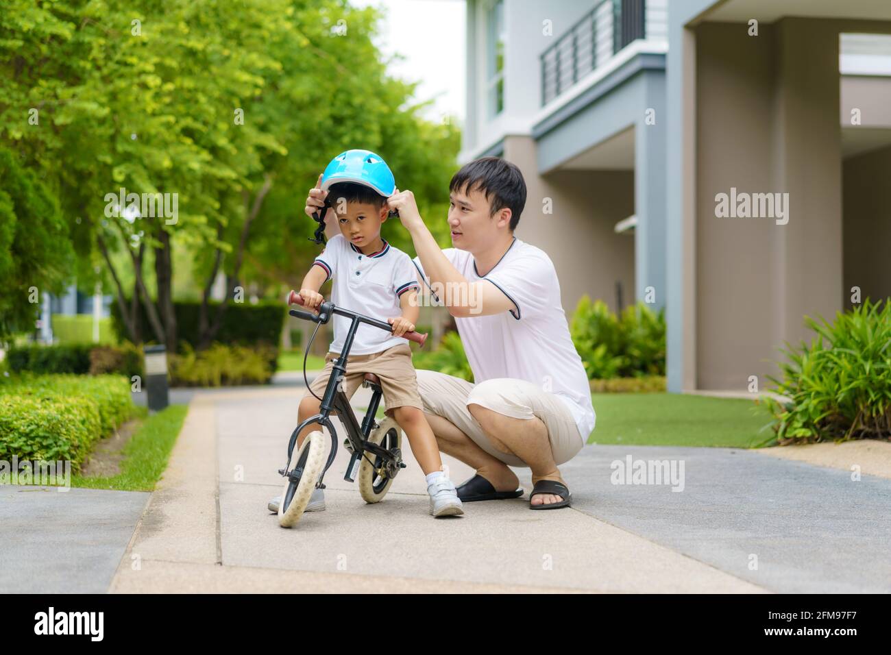 Der asiatische Vater trägt einen Helm für seinen Sohn während er seinem Kind beibringt, wie man in einem Nachbarschaftsgarten Fahrrad fährt, interagieren Väter mit ihren Kindern Stockfoto