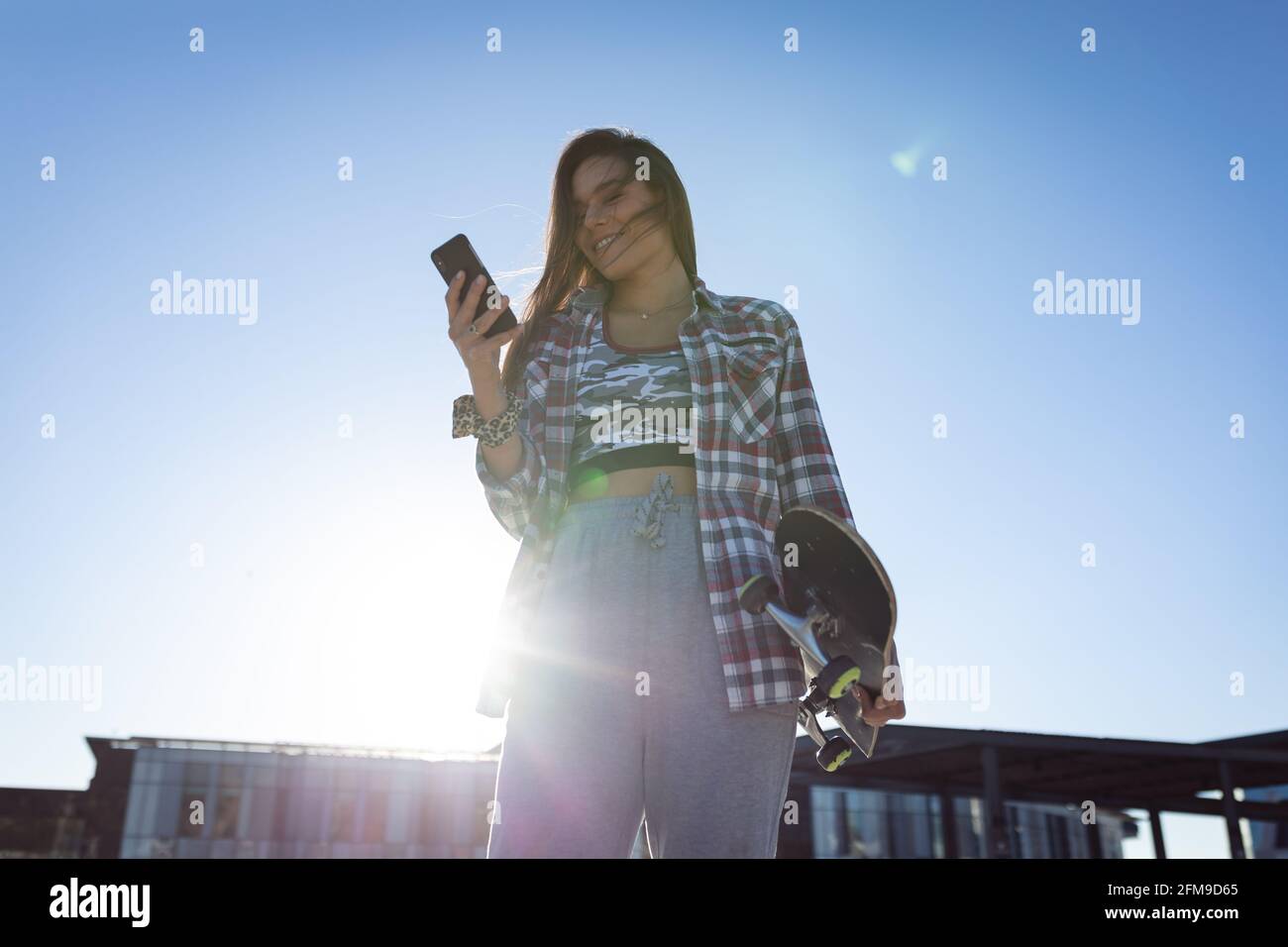 Kaukasische Frau mit Skateboard mit Smartphone in der Sonne Stockfoto