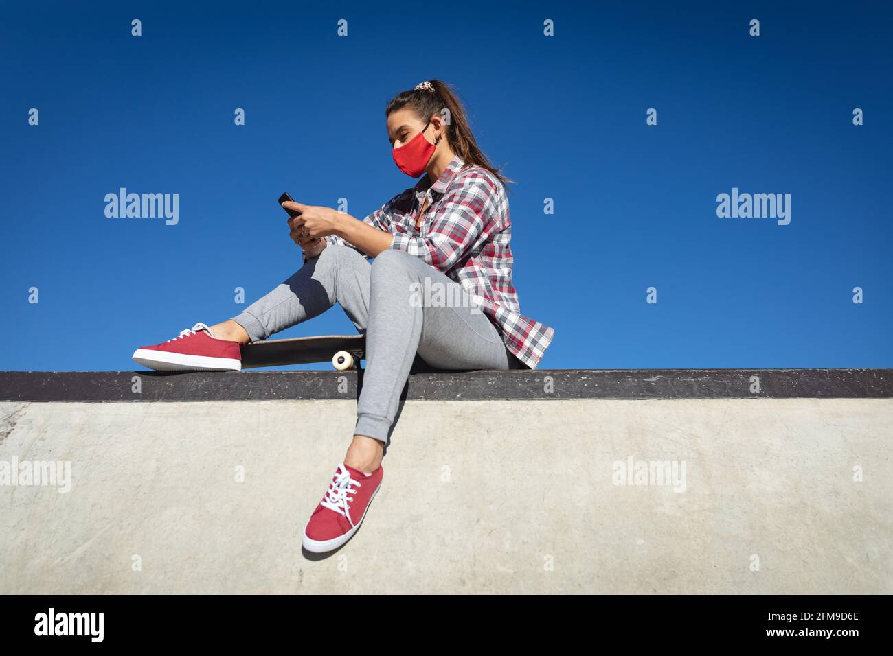 Kaukasische Frau trägt Gesichtsmaske, sitzt an der Wand mit Skateboard und mit Smartphone Stockfoto