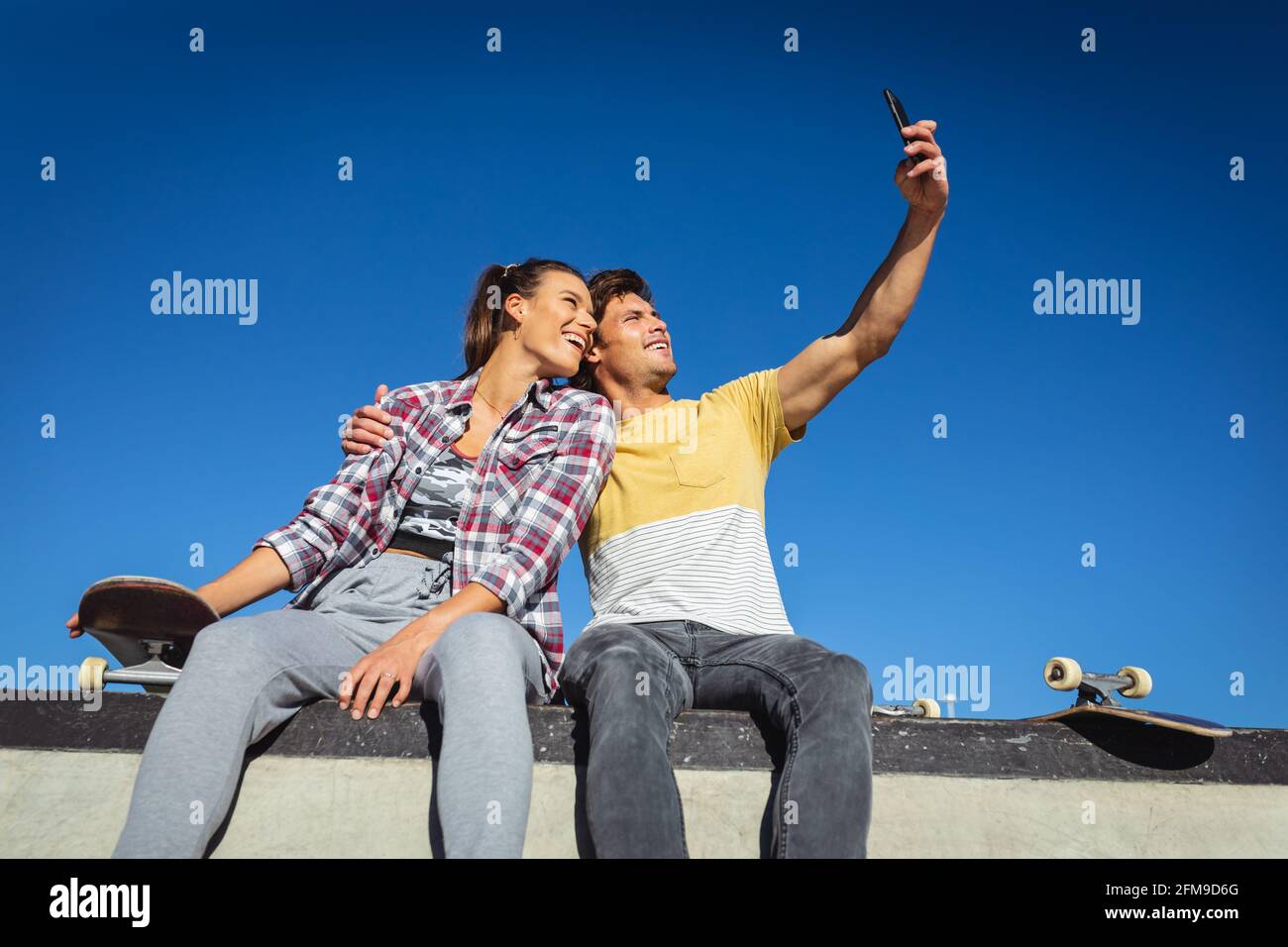 Fröhliche kaukasische Frau und Mann, die mit Skateboards an der Wand sitzen und Selfie in der Sonne machen Stockfoto