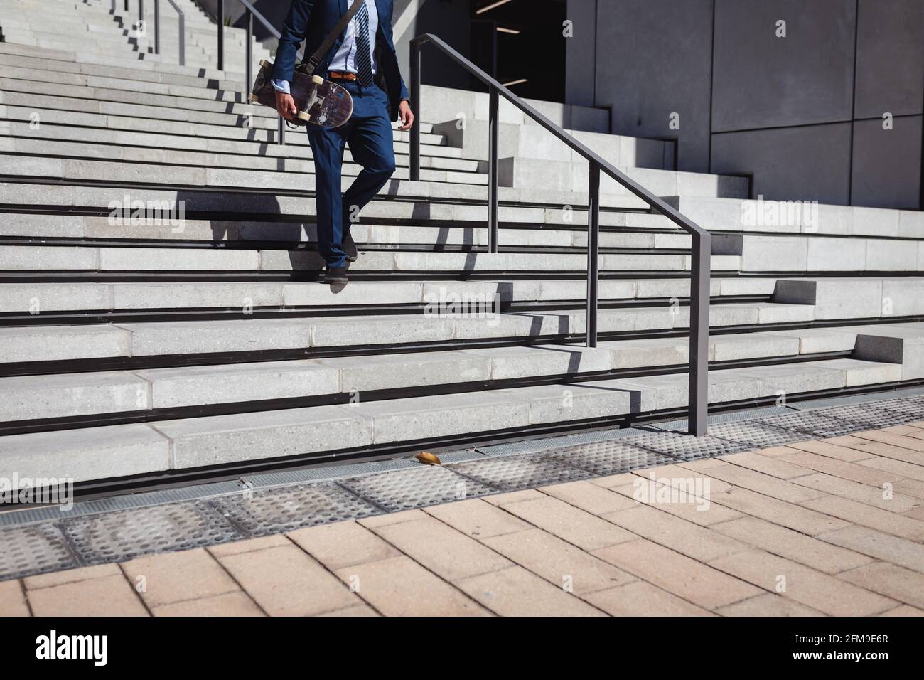 Niedriger Abschnitt des kaukasischen Geschäftsmannes, der Skateboard hält und in der Sonne nach unten geht Stockfoto