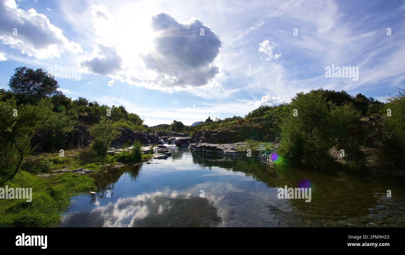 Italien, Sizilien, Alcantara, Alcantara-Schlucht, Fluss, Weitwinkelansicht des Flusses, Hintergrundbeleuchtung, Sonne und Wolken spiegeln sich im Fluss, polierte Felsen im Fluss, grüne Ufer Stockfoto