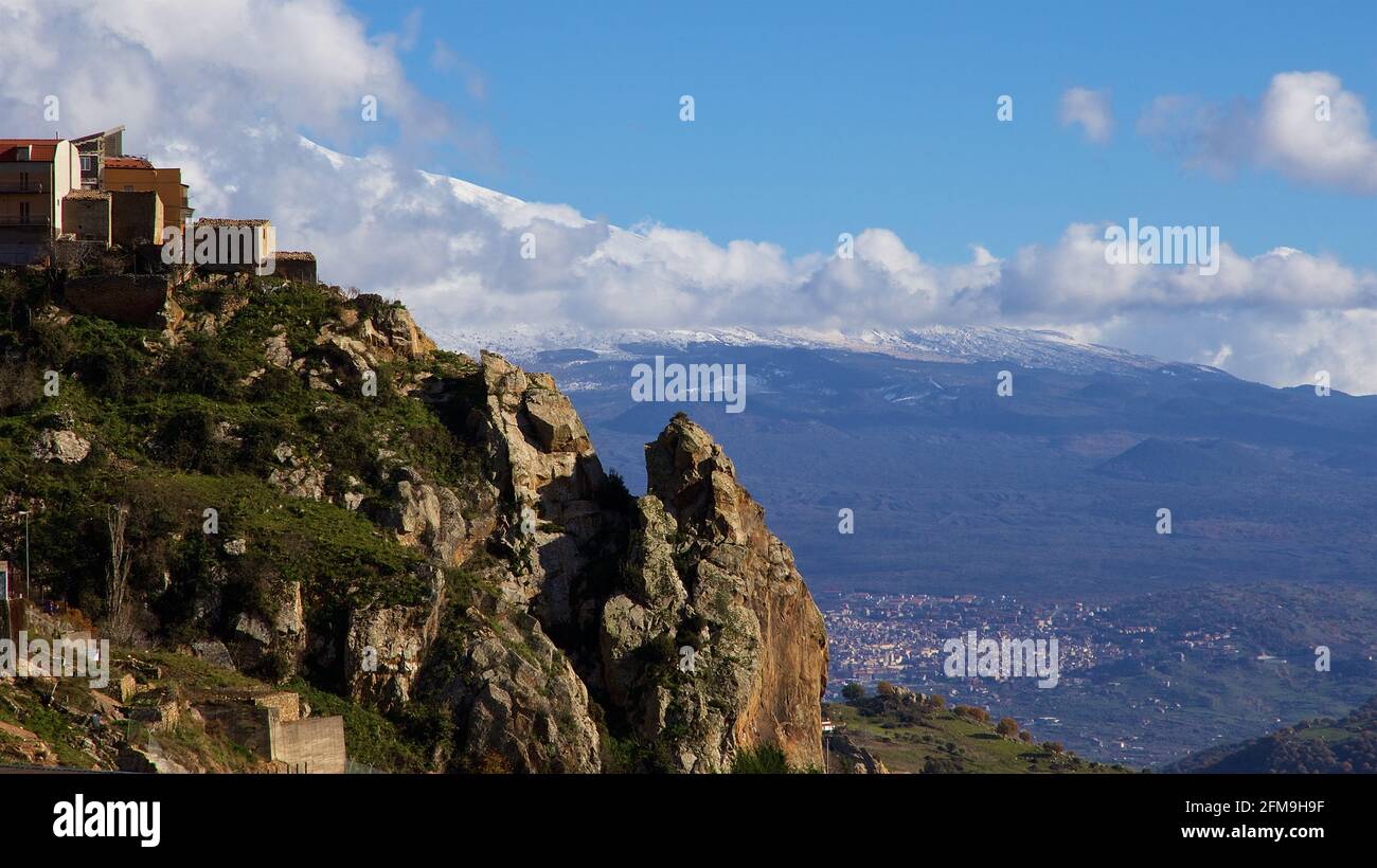 Italien, Sizilien, Blick auf einen Teil des Dorfes Cesaro, auf einem Hügel gelegen, felsigen Vorgebirge, in der mittleren Entfernung unten ist die Stadt Randazzo. Der schneebedeckte Ätna erhebt sich im Hintergrund, der Himmel ist hellblau mit weiß-grauen Wolken Stockfoto