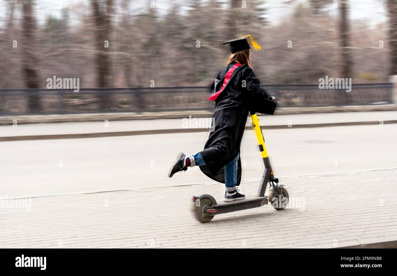 Glückliche junge Frau in schwarzem Abschlusskleid und schwarzer Abschlussfeier Kappe mit gelber Quaste auf einem Elektroroller nach Abschluss der Ausbildung An der Universität o Stockfoto