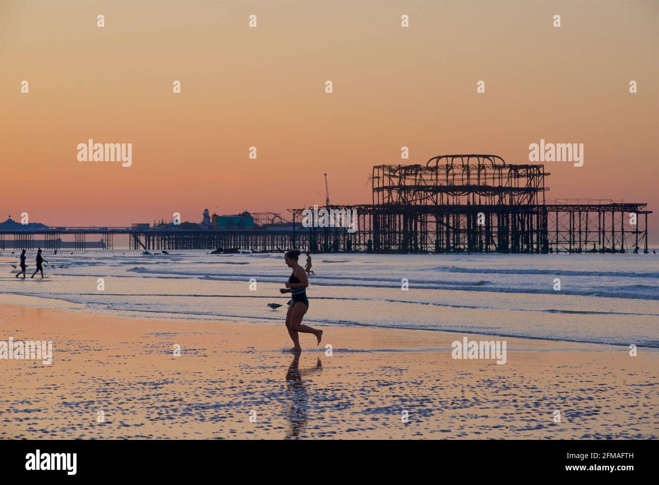 Kommt aus einem frühen Einbruch des Mondes. Schwimmer, der vom englischen Kanal in Brighton aus in die Stadt fährt, mit den silhouettierten Formen der Pfeiler im Hintergrund. Brighton & Hove, Sussex, England, Großbritannien Stockfoto