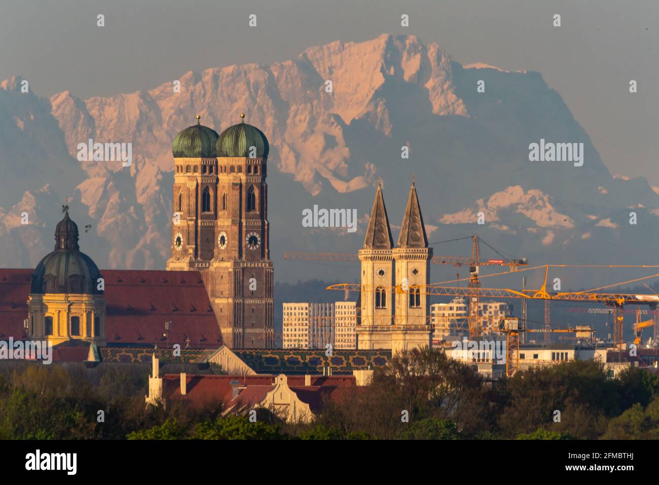 München, Deutschland. Mai 2021. Die Theatinerkirche (l-r), die Frauenkirche und die Sankt Ludwig-Kirche der bayerischen Hauptstadt sind in den frühen Morgenstunden vor der Kulisse der Alpen mit der Zugspitze in etwa 90 Kilometern Entfernung zu sehen. (Das Bild wurde mit einem 1120 Millimeter Teleobjektiv aufgenommen) Quelle: Peter Kneffel/dpa/Alamy Live News Stockfoto