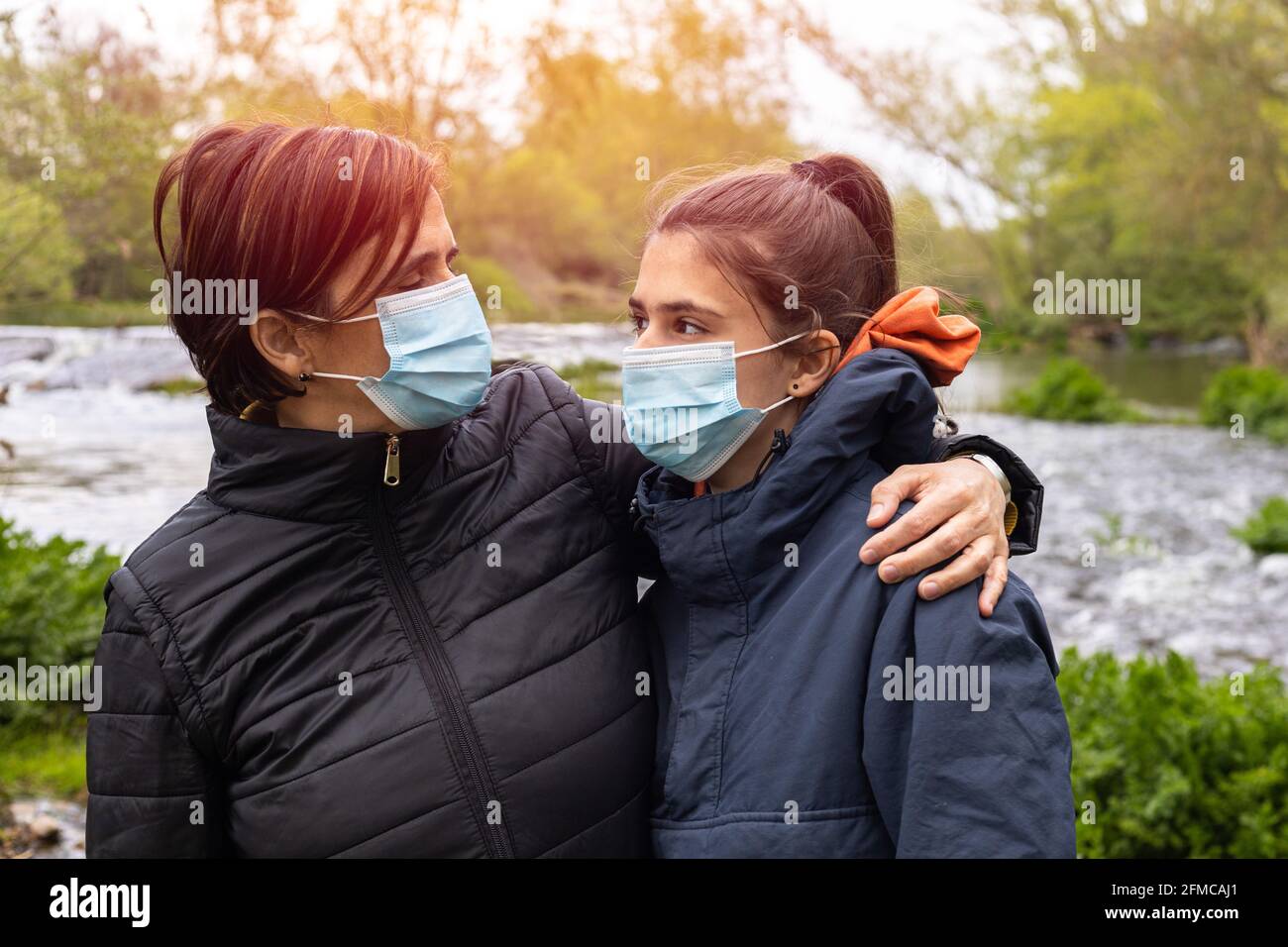 Mutter und Tochter lächeln mit der Coronavirus-Maske in der Feld neben einem Bach bei Dämmerung mit selektivem Fokus Stockfoto