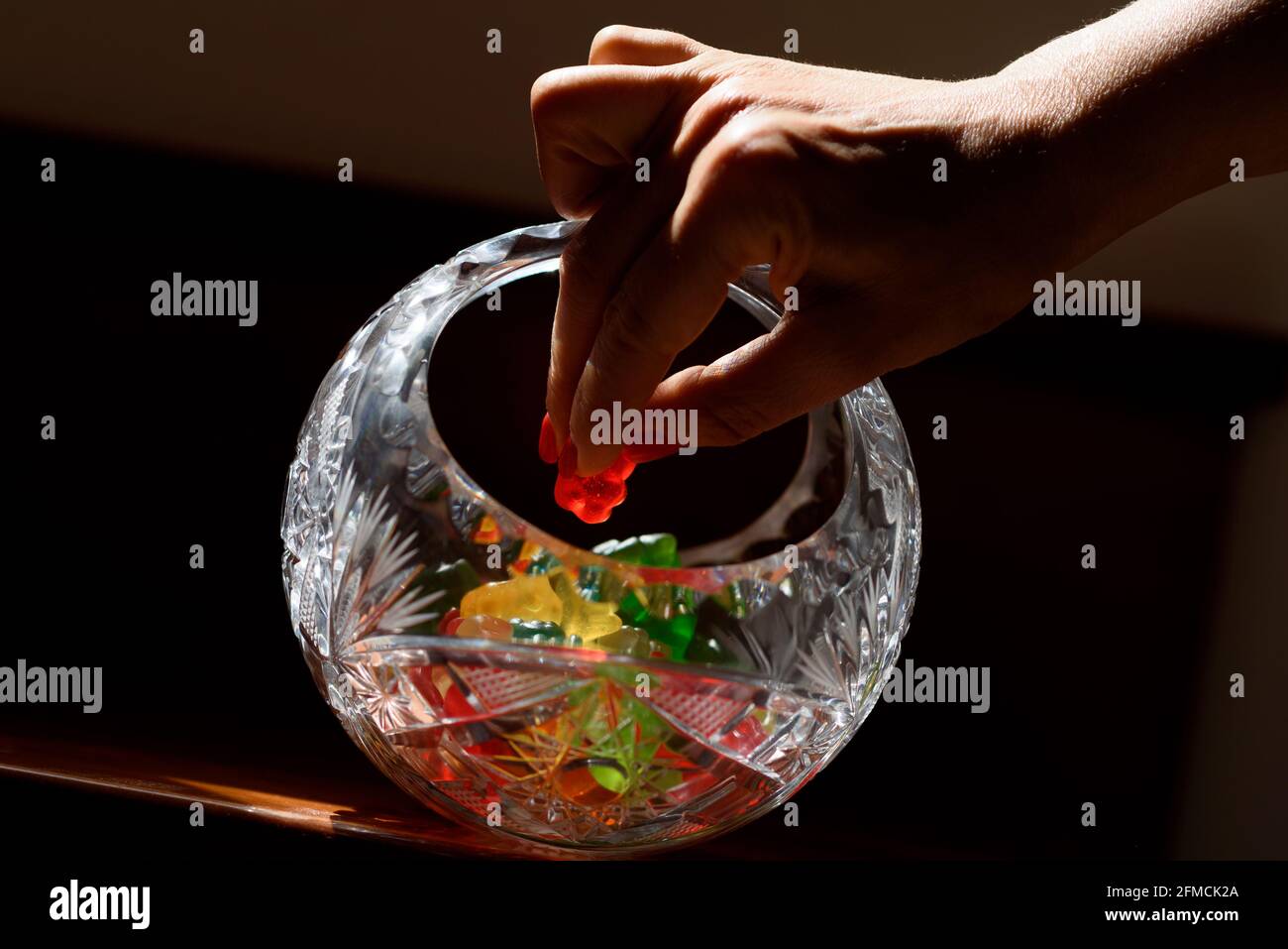 Nahaufnahme einer Frau, die einen roten Gummibären aus einer Kristallvase mit einer Auswahl an bunten Süßigkeiten Gummibären nimmt. Low-Key-Bild. Stockfoto