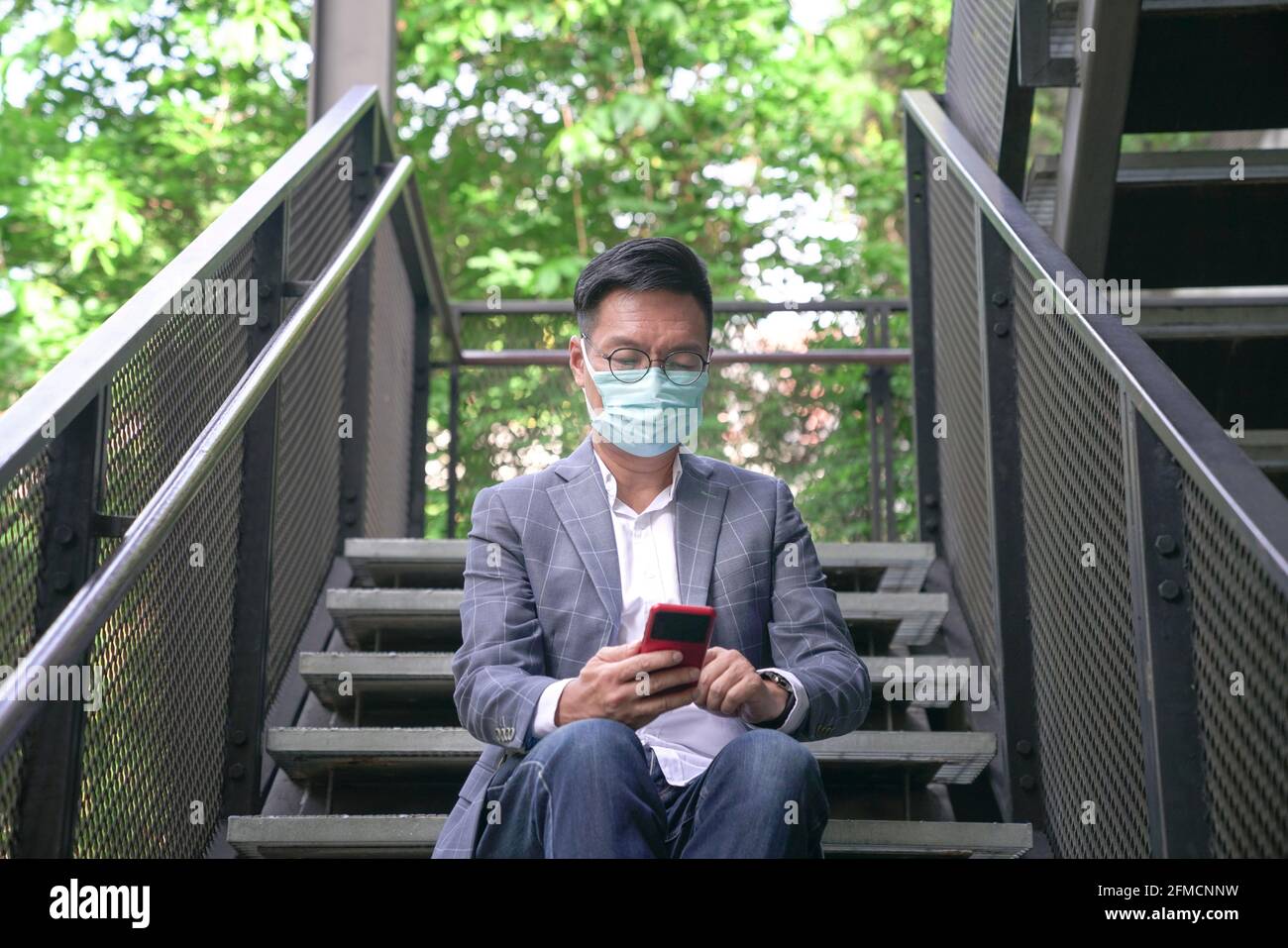 Geschäftsmann mit Gesichtsmaske sitzt auf der Treppe und surft mit seinem Telefon. Urban mit grüner Umgebung. Stockfoto