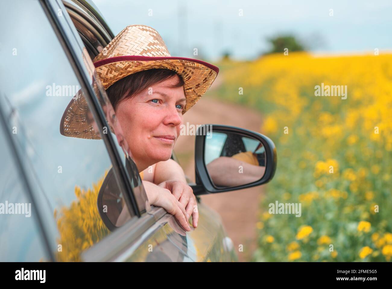 Frau genießt die Autofahrt in blühender Sommerlandschaft, Kopf mit Strohhut, der aus dem Fenster reicht, selektiver Fokus Stockfoto