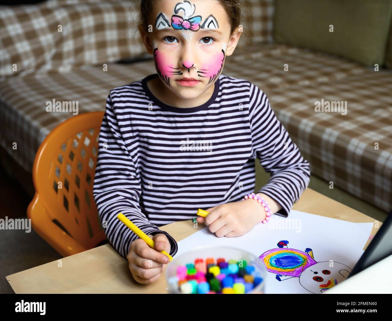 Kinderzeichnung mit Filzstiften innen, kleines Mädchen mit gemalter Maske im Gesicht, das zu Hause studiert. Niedliches Kind lernt am Tisch im Zimmer zu zeichnen. Porträt von Stockfoto