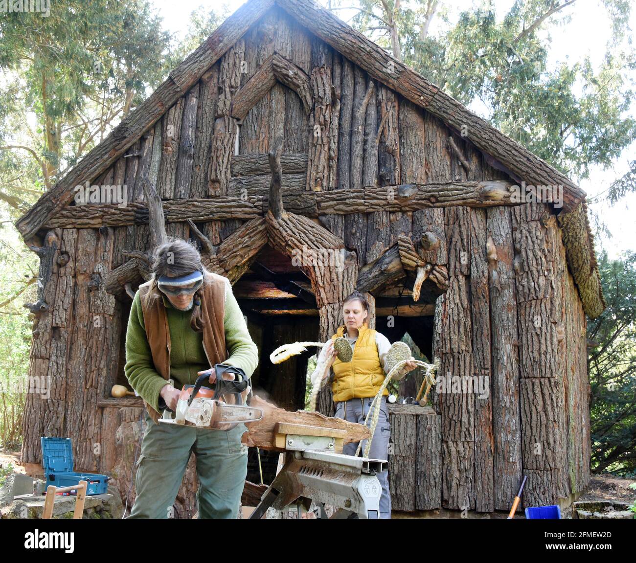 08. Mai 2021, Sachsen-Anhalt, Wörlitz: Die Diplom-Restauratorin Kerstin Klein (r) und die Bildhauerin Katharina Günther bringen die letzten Teile von Plattenbrettern auf die fehlenden Teile auf dem über 230 Jahre alten Borkenhäuschen im Wörlitzer Park auf. Das kleine Holzhaus, auch Wurzelhütte genannt, wurde in den letzten zwei Jahren umfassend restauriert. Die Borkenhäuser dienten Prinz Franz als privates Badehaus zum Umkleiden vor dem Schwimmen im Wörlitzer See und sind außen mit knarrierten Eichenstämmen, Platten und Rinde verkleidet. Innen schmücken Kassen aus Ulmenhalbstrümmen und mit handgewebten Schilfmatten und Rush-Zöpfen die Stockfoto