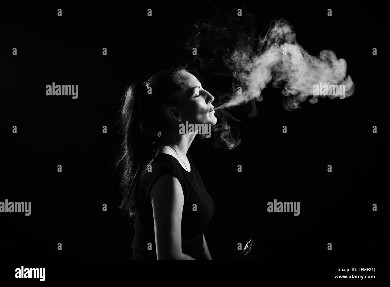 Die Brünette raucht auf schwarzem Hintergrund eine elektronische Zigarette und gibt dicken Rauch aus ihrem Mund frei. Monochrom. Porträt einer Frau, die einen Vape raucht Stockfoto