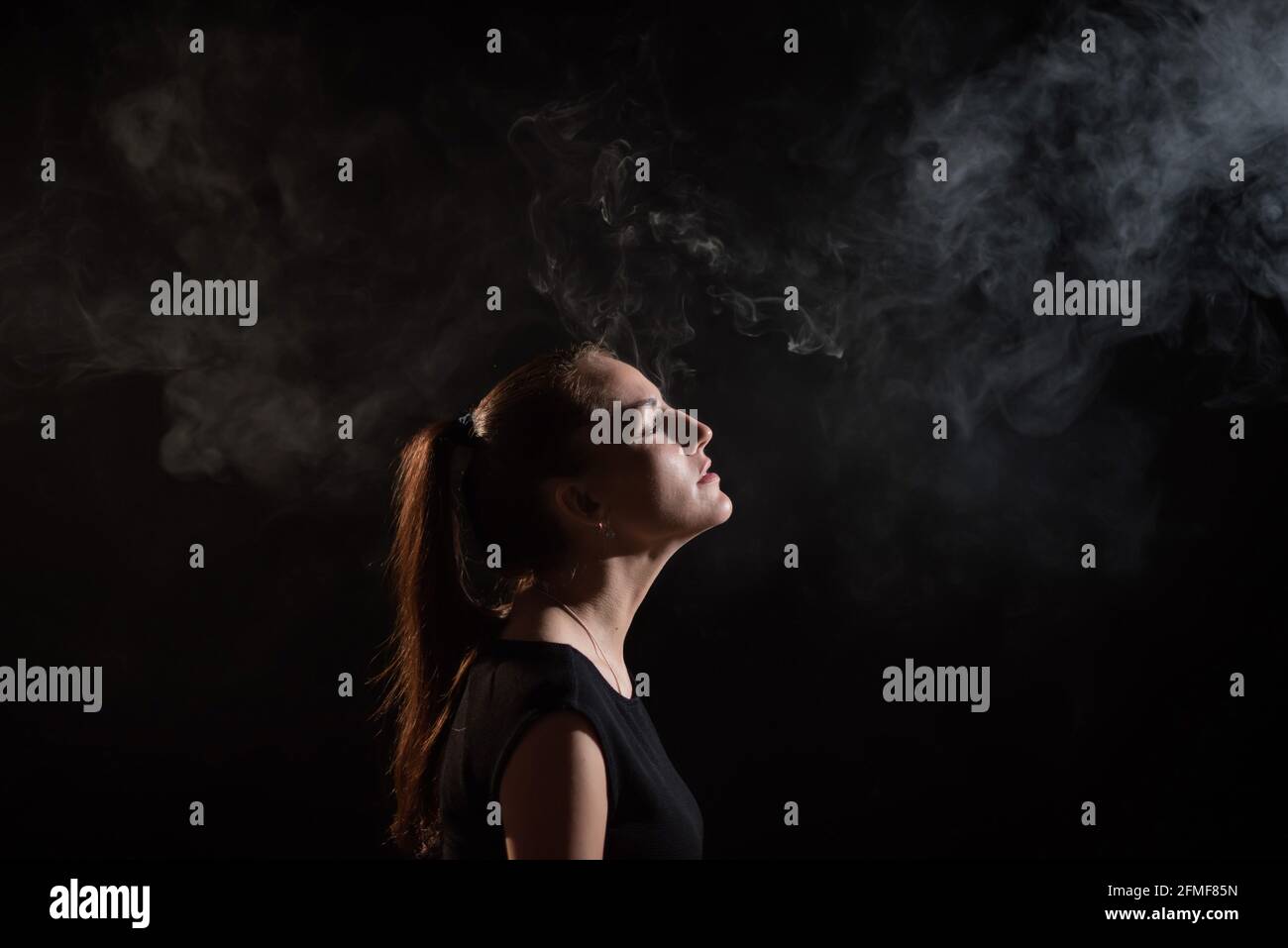 Die Brünette raucht auf schwarzem Hintergrund eine elektronische Zigarette und gibt dicken Rauch aus ihrem Mund frei. Porträt einer Frau, die einen Wape raucht, schwebend. Stockfoto
