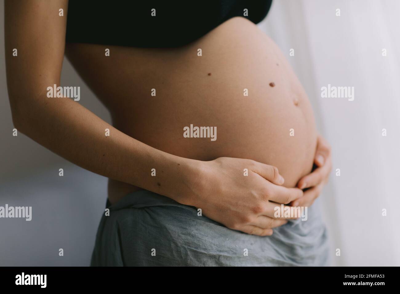 Eine Schwangerin berührt ihren Bauch in der Nähe der Fenster. Schwangerschaft und Mutterschaft Konzept. Stockfoto