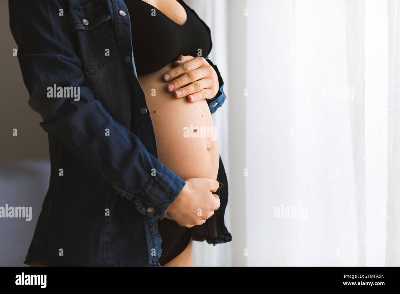 Eine Schwangerin berührt ihren Bauch in der Nähe der Fenster. Schwangerschaft und Mutterschaft Konzept. Stockfoto