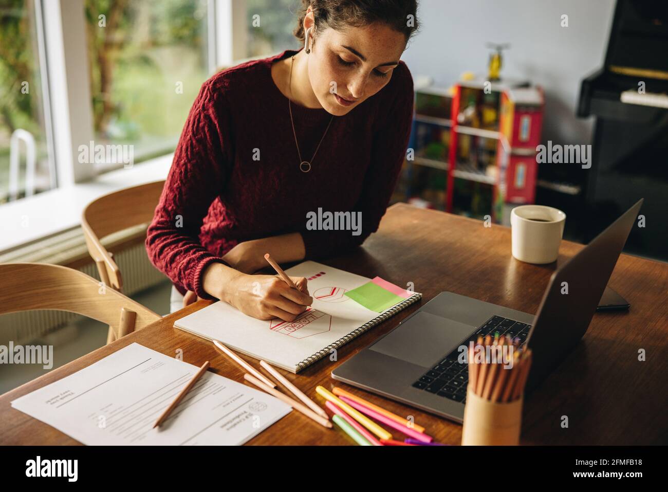 Schöne Frau Illustratorin Zeichnung auf Papier. Eine Künstlerin sitzt am Tisch mit einem Laptop und macht zu Hause einige Illustrationen mit einem Farbstift. Stockfoto