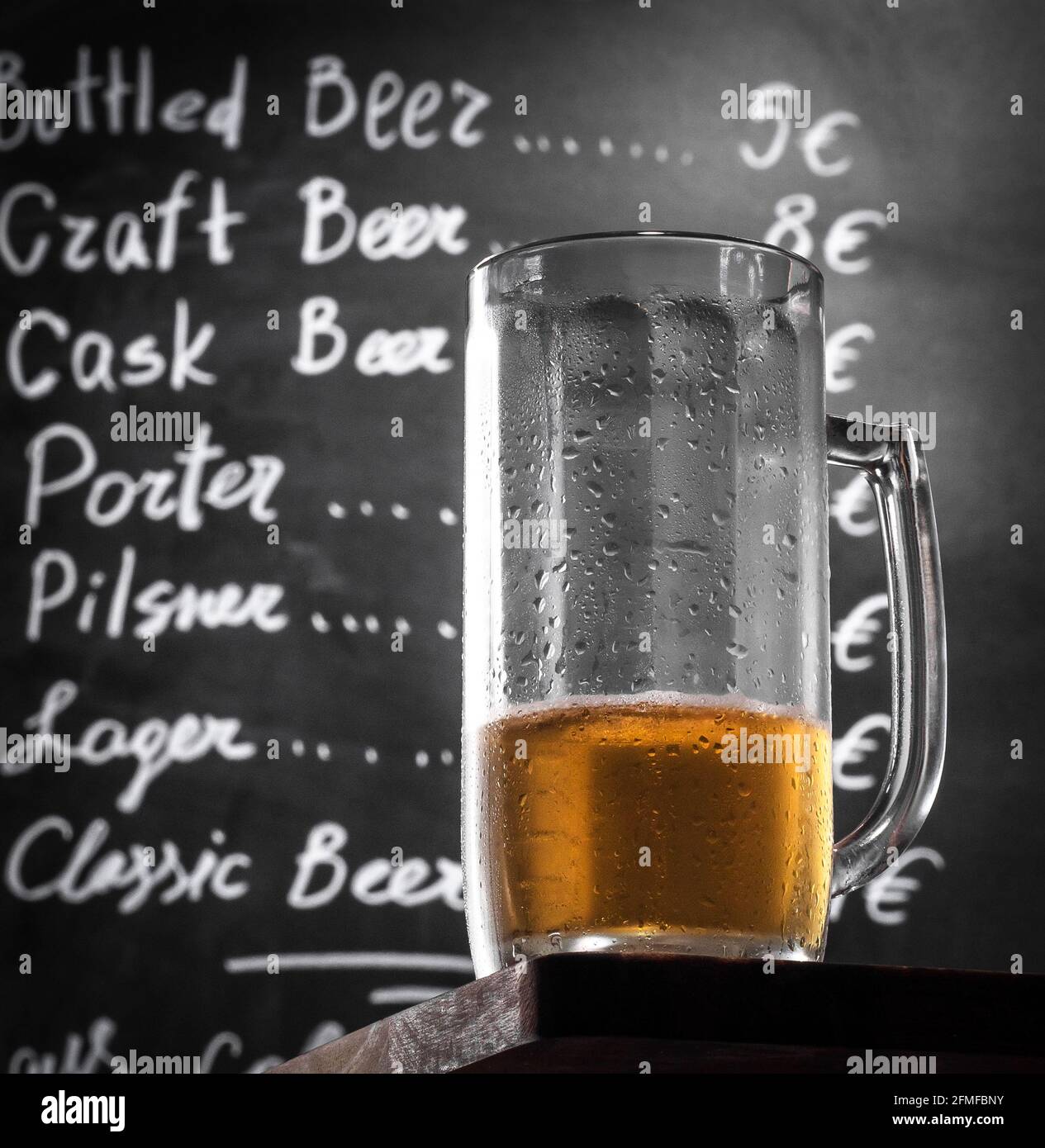 Becher Bier ohne Schaum auf einem Holztisch an der Kreidetafel mit Barmenü. Unfertiges Glas eines kalten Fassbiers auf einem Pub-Tisch, Nahaufnahme. Stockfoto