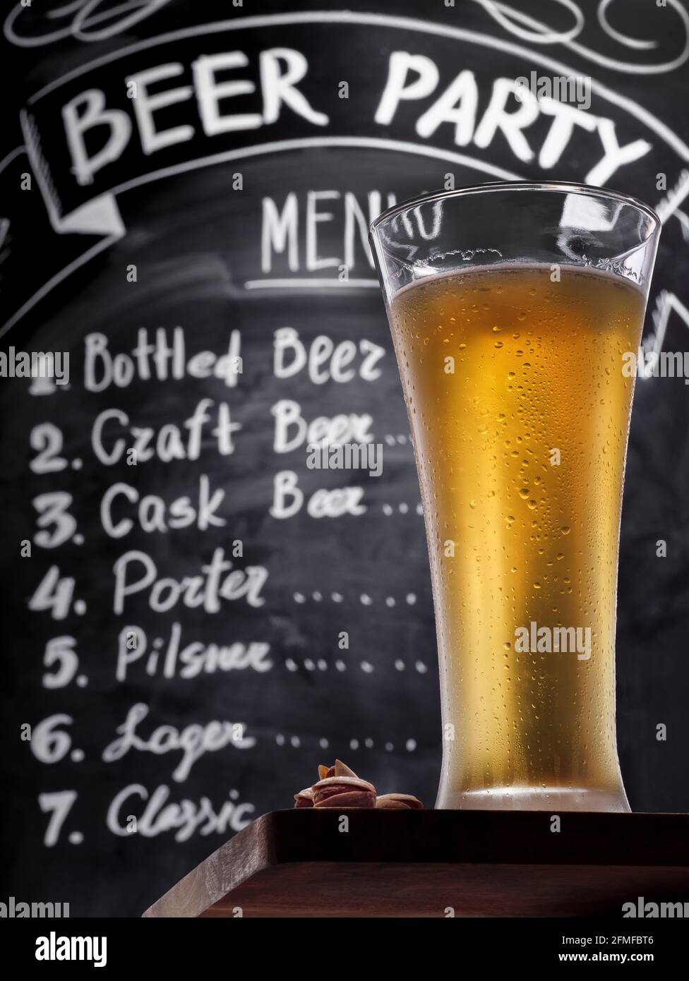 Volles Glas kaltleichtes Bier mit Pistazien auf einem Holztisch vor dem Hintergrund einer Kreidetafel mit Menütext, Ansicht von unten nach oben. Bier vom Fass konisch Stockfoto