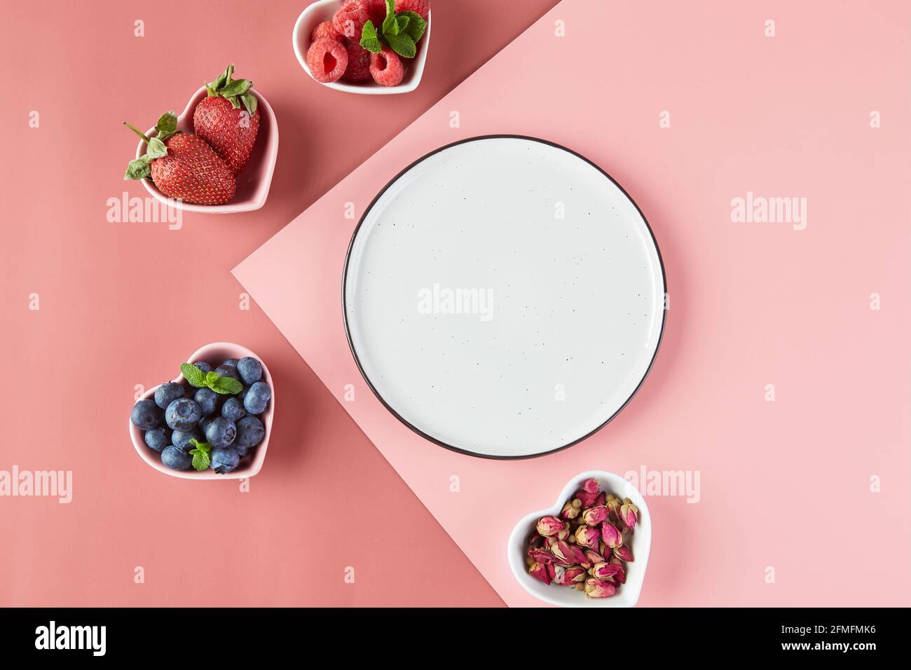 Leere Teller und kleine Teller in Form von Herzen mit frischen Erdbeeren, Himbeeren, Heidelbeeren auf einem rosa Hintergrund. Draufsicht. Stockfoto