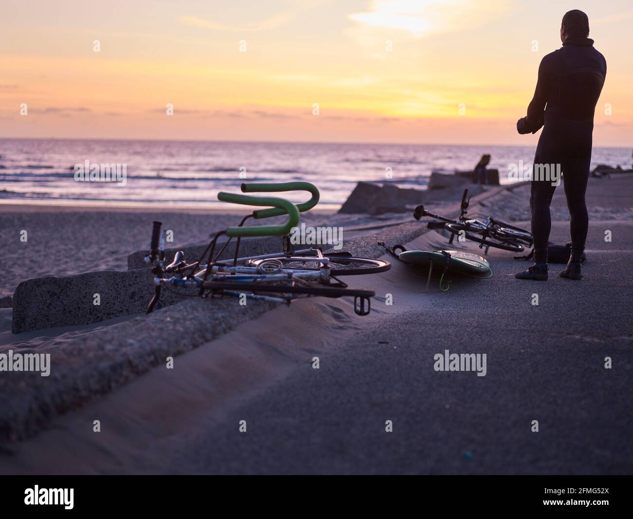 Der Surfer schaut während des Sonnenuntergangs zum Meer, während das Fahrrad zum Transport des Surfbretts neben ihm liegt. Stockfoto