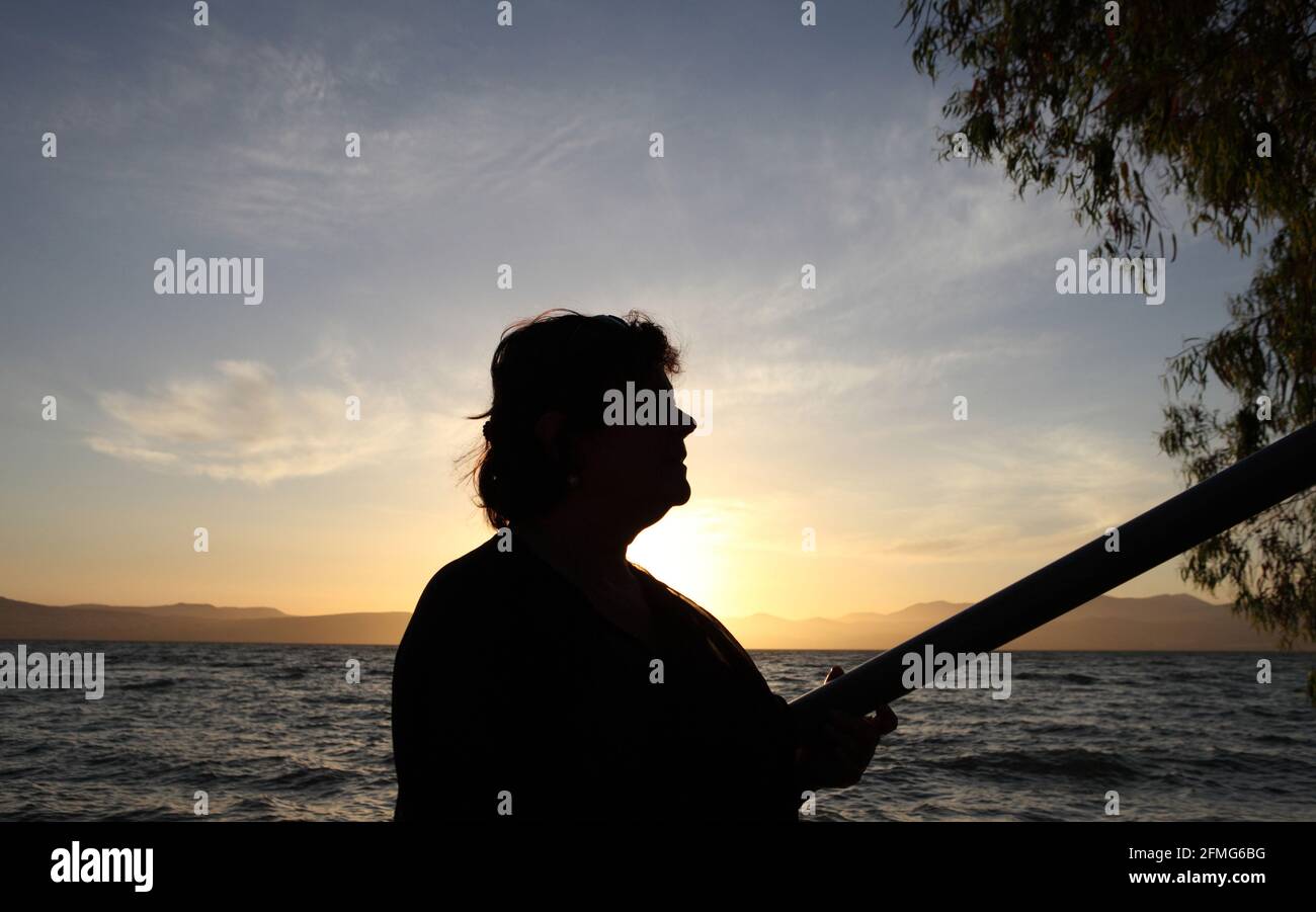 Silhouette bei Sonnenuntergang einer 62-jährigen Frau, der See von Galilee, die Unteren Galiläischen Berge und der bunte wolkige Himmel über ihnen sind hinter. Stockfoto