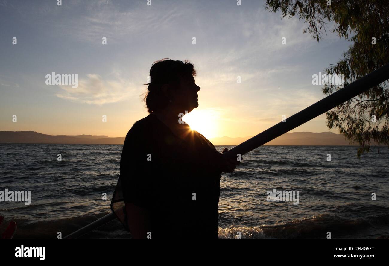 Silhouette bei Sonnenuntergang einer 62-jährigen Frau und Sonnenstrahlen, der See von Galilee, die Unteren Galiläischen Berge und ein dramatischer wolkiger Himmel sind hinter sich. Stockfoto