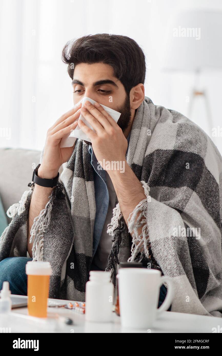 Bleiben Sie zu Hause, wenn Sie sich krank fühlen. Kranker araber, der mit einer Decke bedeckt war und seine Nase niest, auf dem Sofa im Wohnzimmer sitzend, abgeschnitten Stockfoto