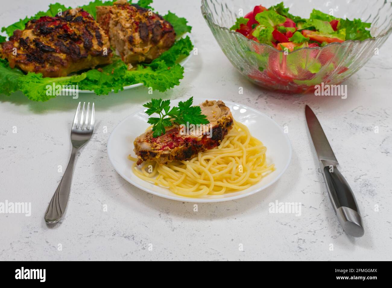 Ein Teller Pasta und ein großes Stück Schweinefleisch, im Ofen gekocht, im Ofen gebacken. Im Ofen gebackenes Fleisch auf grünen Salatblättern. Mit Pasta auf einem Teller mit Stockfoto