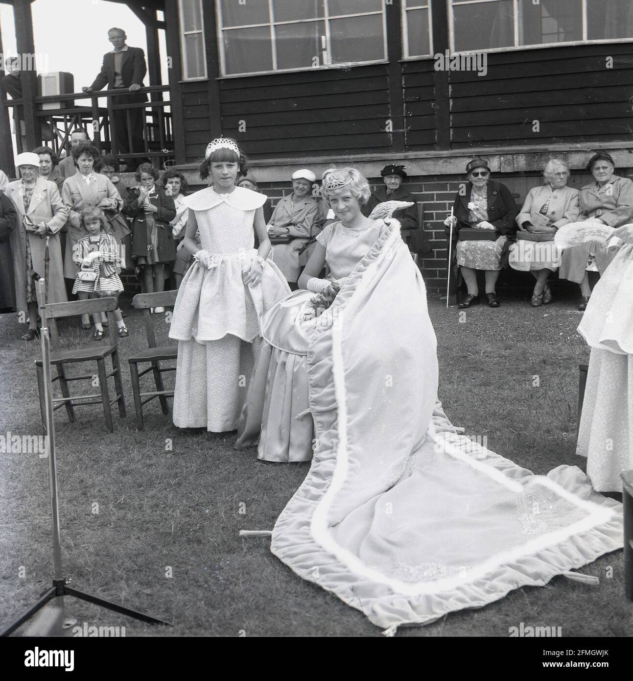 Die 50er Jahre, historisch, draußen in einem Park, beobachten Einheimische die May Queen der Stadt, die auf einem Stuhl auf dem Gras sitzt, mit einem langen Zug ihres Kleides auf dem Boden, Farnworth, Lancashire, England, Großbritannien. Stockfoto