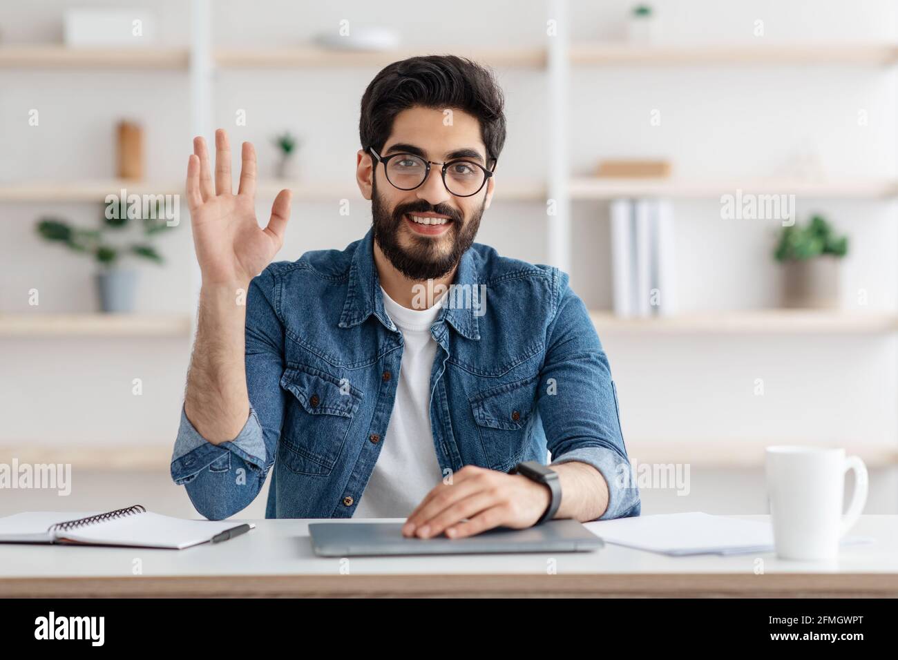 Glücklicher araber, der mit der Hand winkt und lächelt zur Kamera und zu Hause am Arbeitsplatz sitzt. Modernes Fernkommunikationskonzept Stockfoto