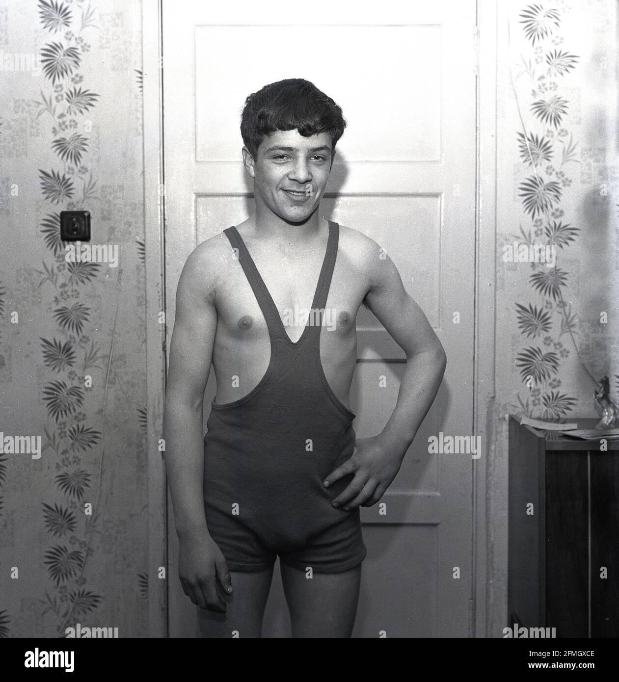 1960s, historisch, ein junger männlicher Wrestler, der in seinem Wrestling Singlet oder einfach Singlet steht, Kelty, Schottland, Großbritannien. Das Singlet ist ein einteiliges, eng anliegender Kostüm oder Sportkit, das den Körper von der Mitte der Brust aus bedeckte, aber die obere Körperhälfte und die Arme ohne Abdeckung frei ließ, Obwohl das einteilige Singlet als die traditionelle Uniform des Sports bekannt ist, wurde es in der Tat erst seit den späten 50er Jahren weit verbreitet, mit Wrestlern vor damals, die Hosen über Leggings mit oder ohne Weste trugen, während des Wettkampfs. Wrestling war eine beliebte Aktivität bei den Highland Games. Stockfoto