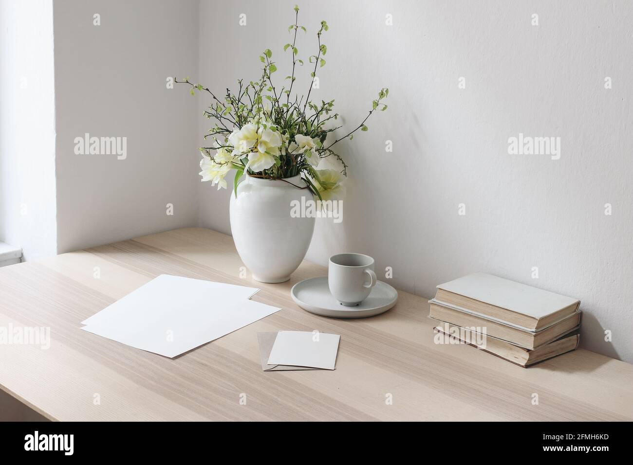 Leere Papierblätter, Grußkarten auf Holztisch. Bücher und eine Tasse Kaffee. Schreibwaren-Modelle. Vase mit gelben Tulpen und Narzissen. Elegant Stockfoto