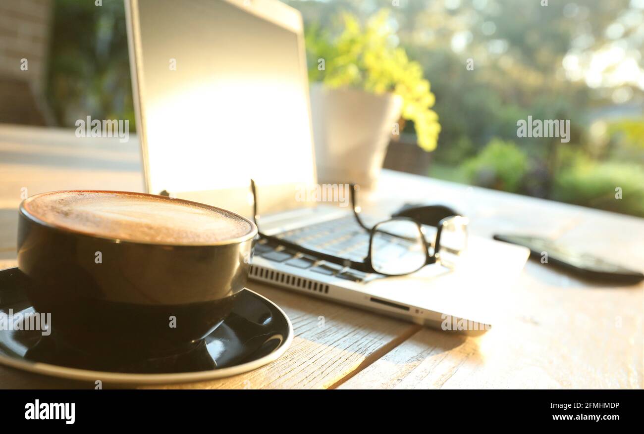 Schöner goldener Morgensonne auf einem Tisch im Freien mit heißem Kaffee, einem Laptop, Handy, Brille und Maus für Online-Sh Stockfoto