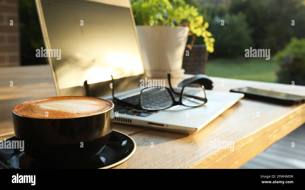 Wunderschöner, fauler, goldener Morgensonne auf einem Tisch im Freien mit heißem Kaffee, einem Laptop, Handy, Gläsern und einer Maus für Onli Stockfoto
