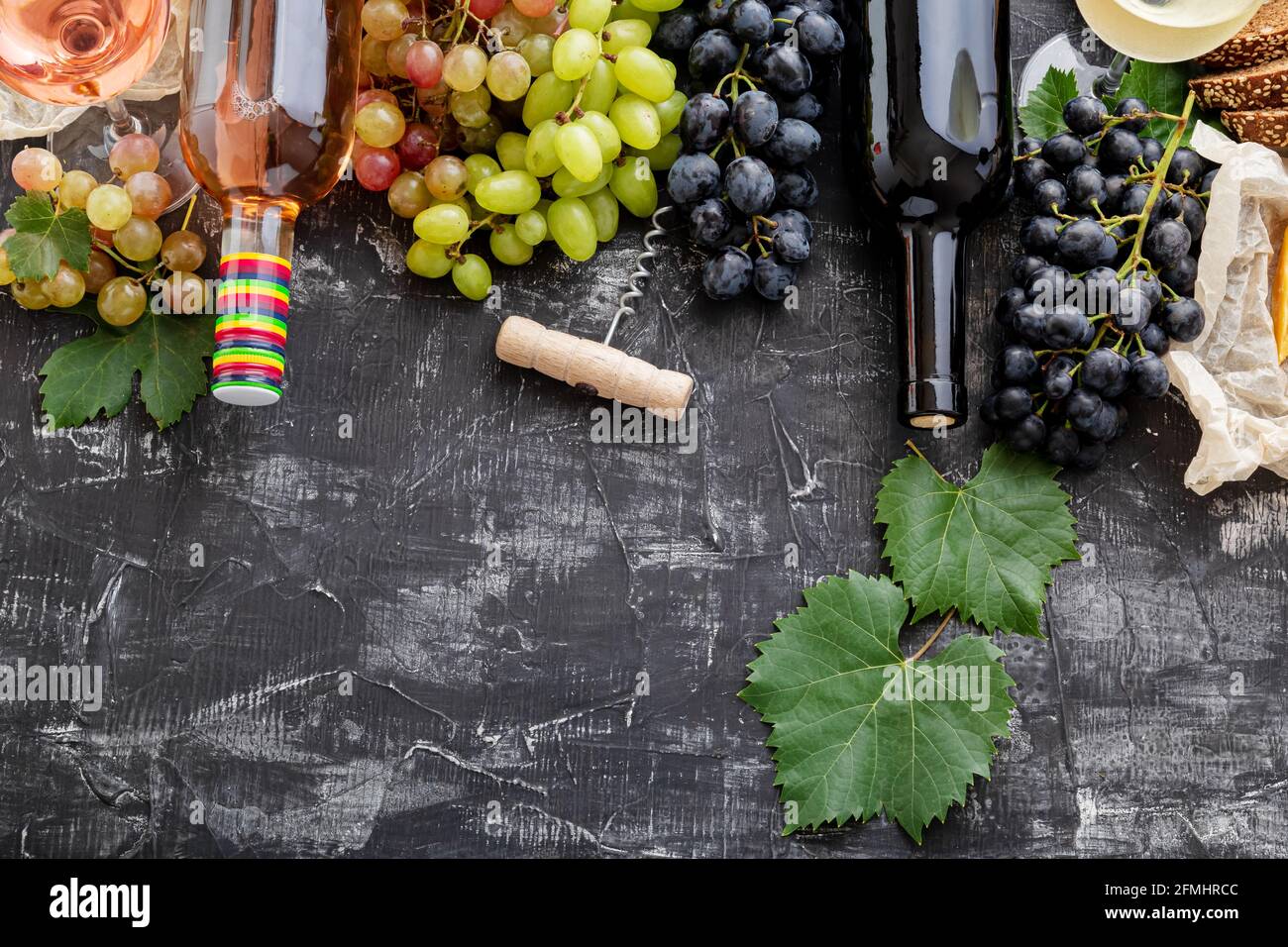 Rosé-Rotwein in Flaschen Käse, weiße rosa und schwarze Trauben mit Weinrebe Pflanze auf dunklem Beton Hintergrund. Sortiment verschiedene Arten von Wein und Trauben Stockfoto