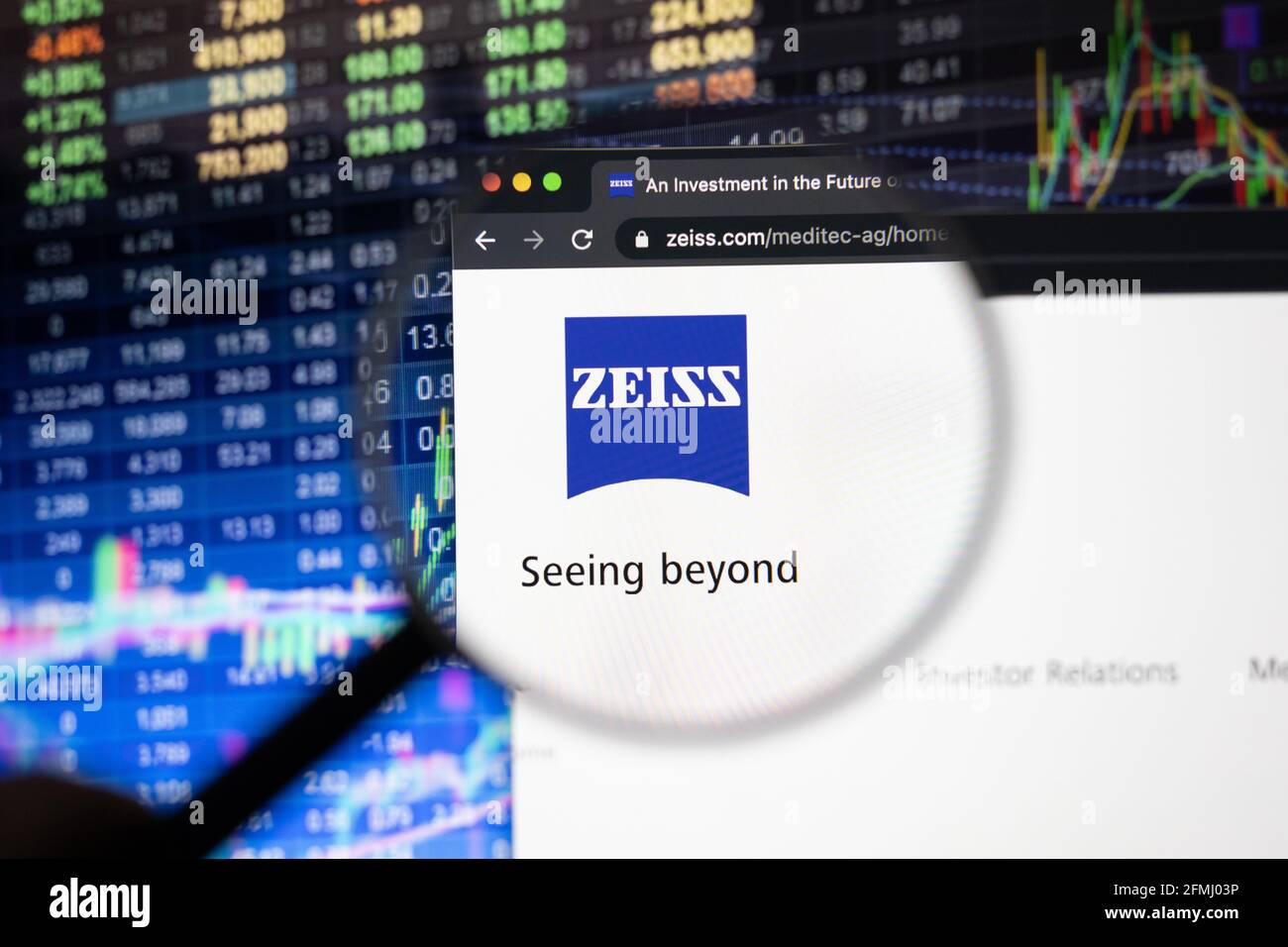 Zeiss Firmenlogo auf einer Website mit verschwommenen Börsenentwicklungen im Hintergrund, die auf einem Computerbildschirm durch eine Lupe gesehen werden Stockfoto